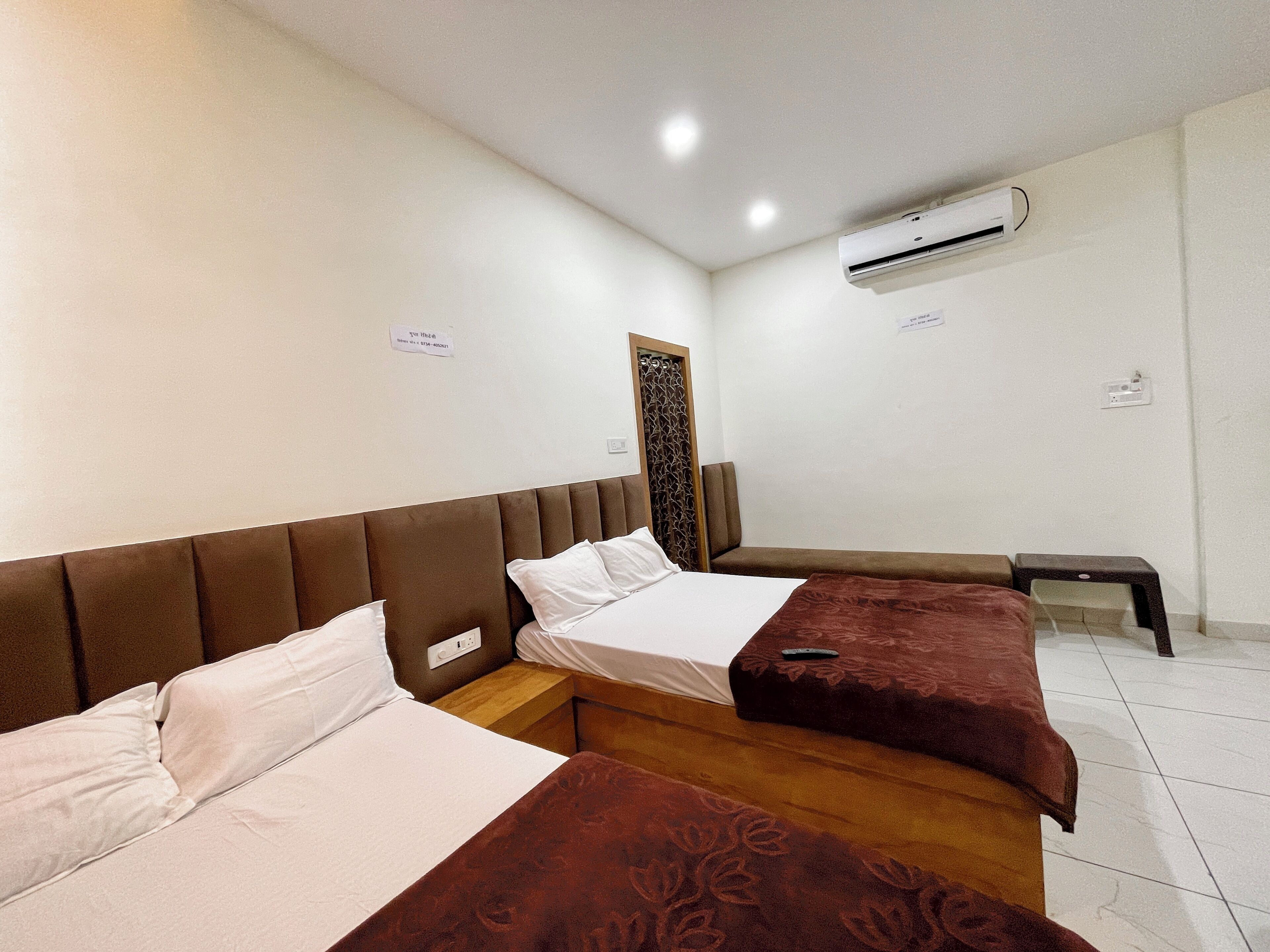 A hotel room with two beds, a wooden headboard, a small bench, an air conditioning unit, and a decorative wall panel.