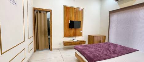Une chambre d’hôtel moderne avec une armoire en bois, un téléviseur à écran plat fixé sur un panneau en bois, une table de chevet avec une lampe et un grand lit recouvert d’une couverture violette.