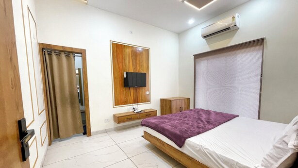A modern bedroom with a wooden bed frame, a flat-screen TV mounted on a wooden panel, a bedside table, and a window with blinds. - Hotel Gupta Residency - near Temple  (Ujjain)