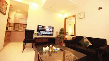 A living room with a flat-screen TV, a black sofa, a glass coffee table, and a kitchen in the background.