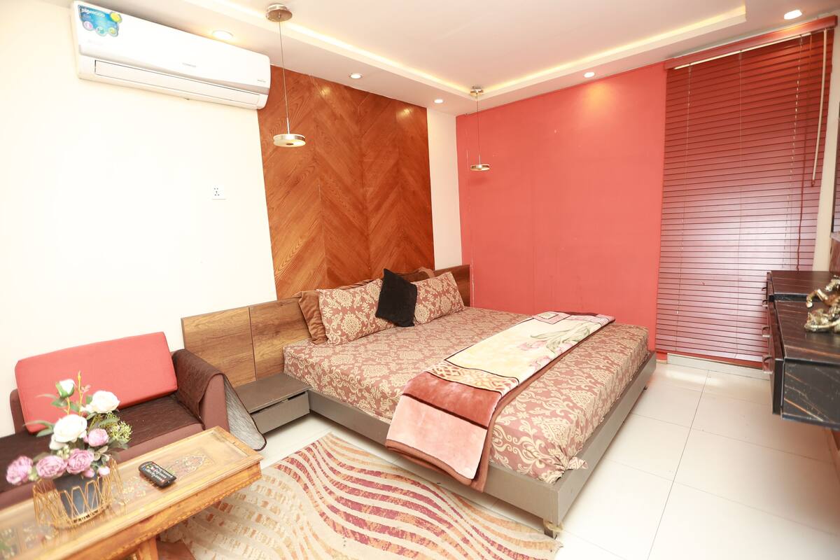 Une chambre à coucher avec un lit, une tête de lit en bois, un mur rouge, une fenêtre avec des stores, une table de chevet ornée d’un vase de fleurs et un petit bureau avec une lampe.