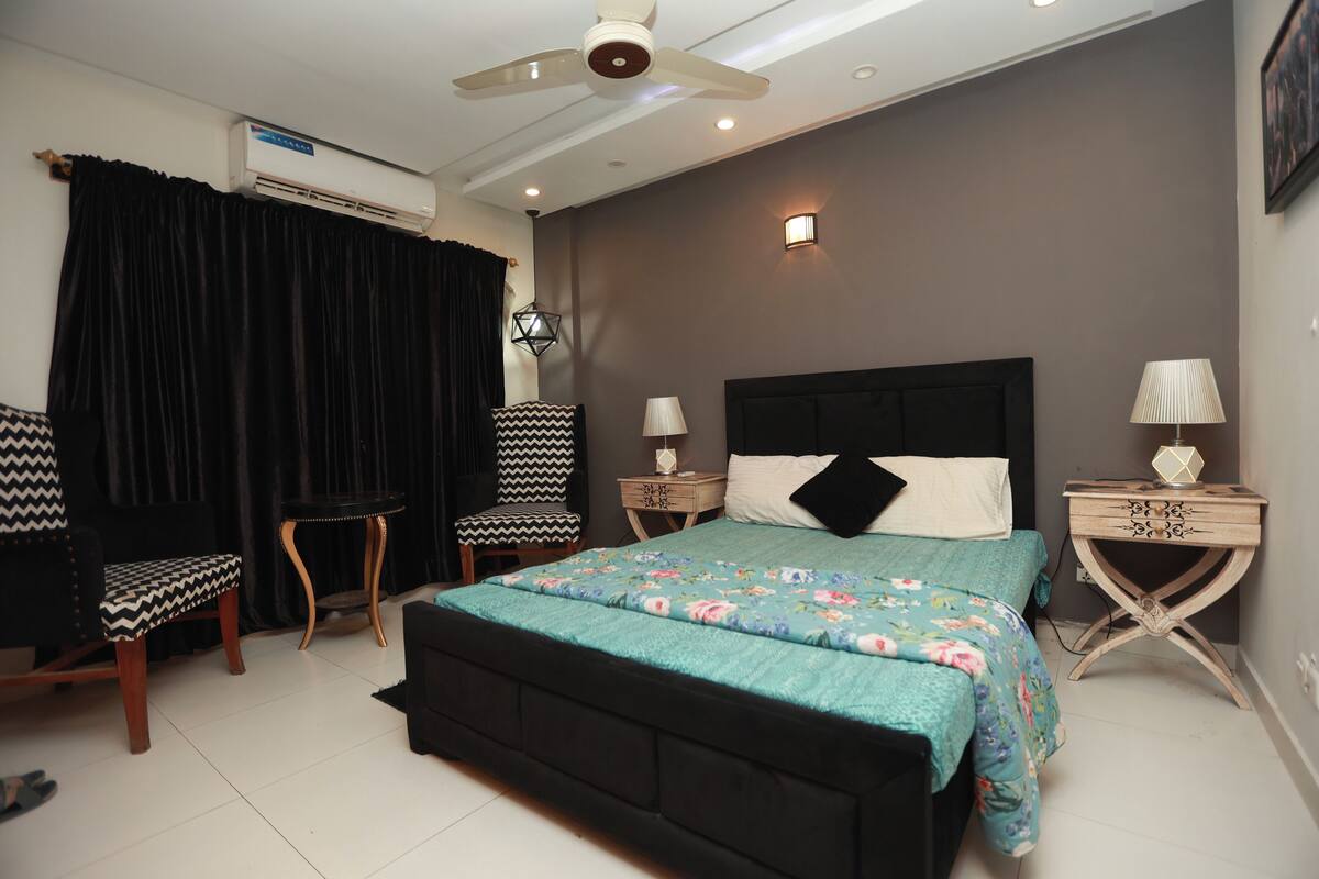 Une chambre à coucher comprenant un lit, deux tables de chevet, une chaise, un ventilateur de plafond et un appareil de climatisation.