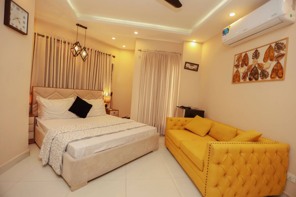 Une chambre à coucher avec un lit, un canapé jaune, une horloge murale et un climatiseur.