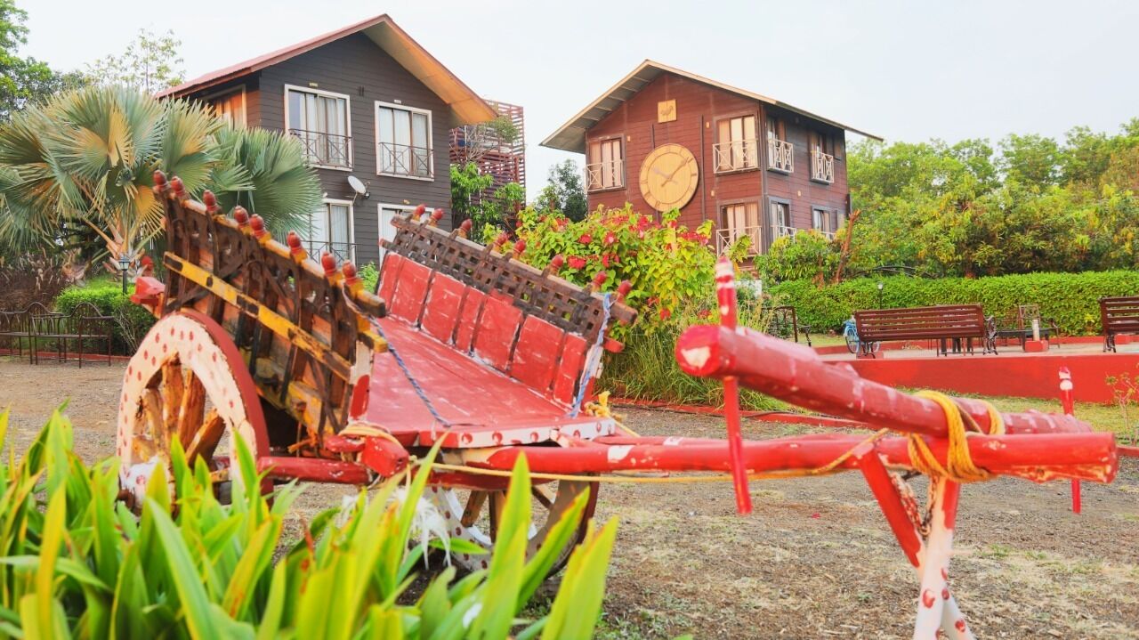 A rustic wooden cart with a red seat, situated in a garden area with two houses in the background.