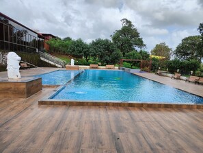 A rectangular swimming pool with a wooden deck surrounding it, benches along the edge, and a statue of a lion. - VITS Select Jayshin Igatpuri (Trimbak)