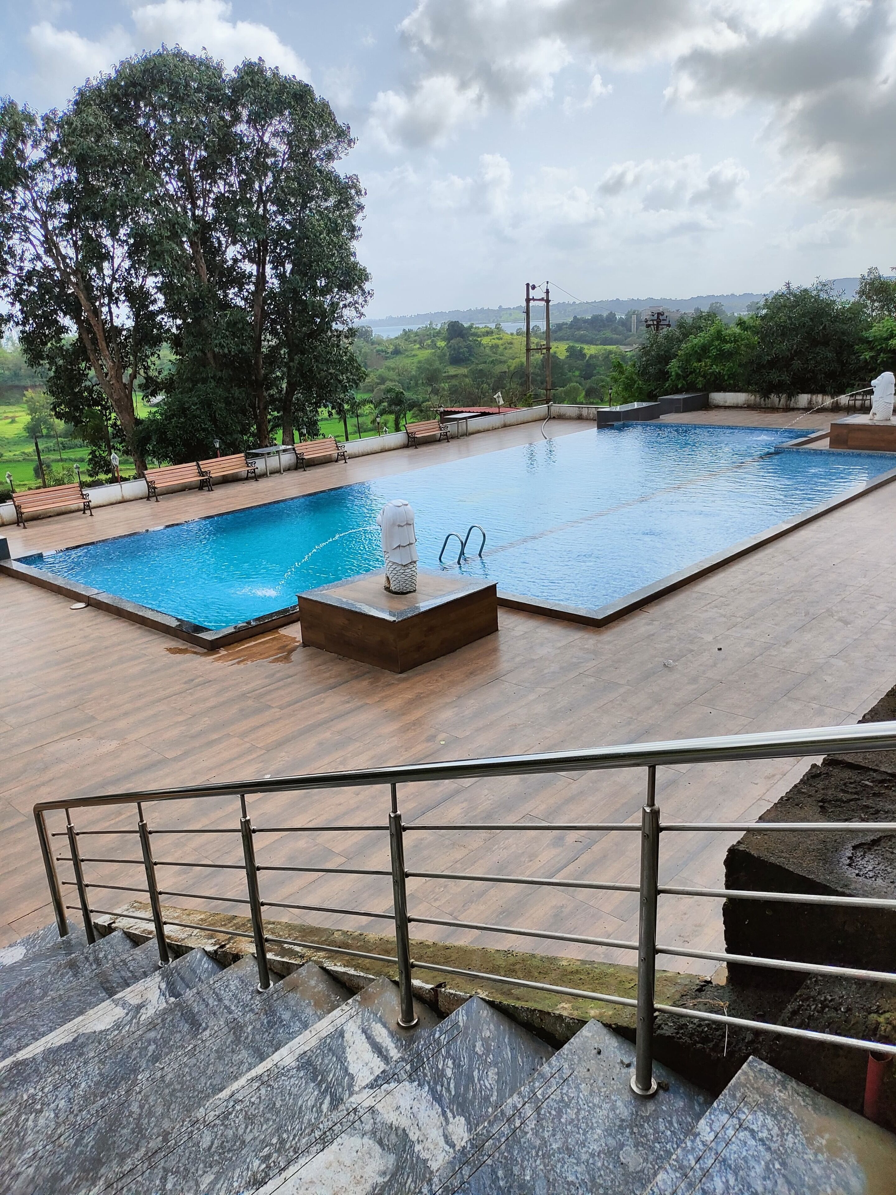 A swimming pool with a view of a green landscape, surrounded by wooden benches and a stone statue.