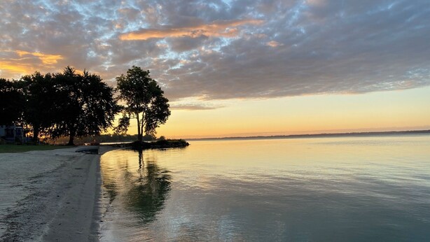 Una playa con aguas tranquilas que reflejan el cielo, árboles y una costa distante.