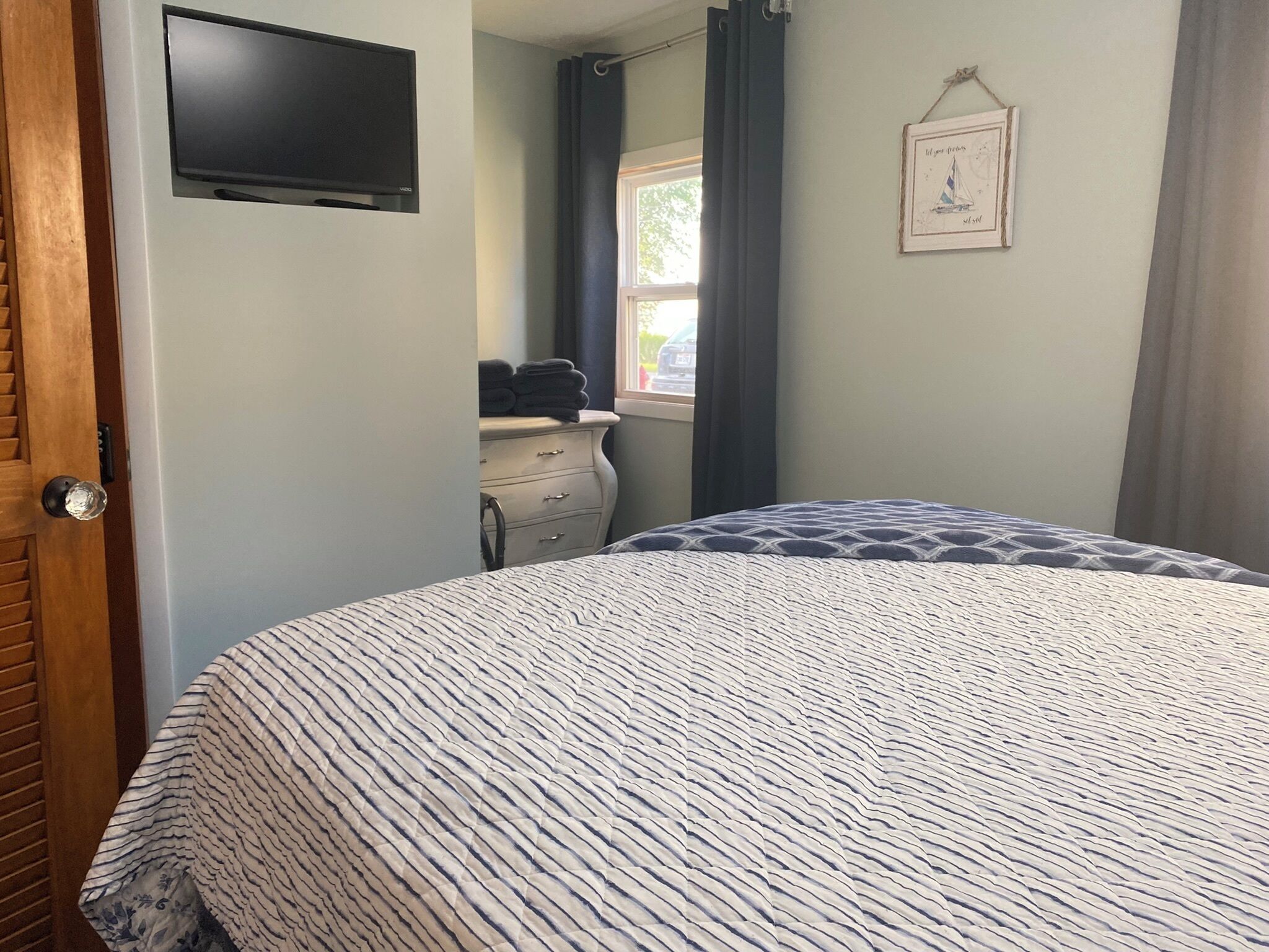 A bedroom with a bed, a dresser, a TV mounted on the wall, and a window with curtains.