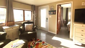 Una habitación bien iluminada con un ventanal grande, una silla con cojín, una mesita, un televisor y un aparador.
