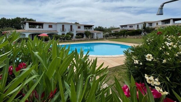 A swimming pool surrounded by lush greenery and white buildings with red accents. - Luisa Apartment In Budoni Shared Pool (Budoni)