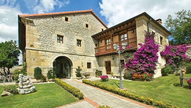 A stone building with a large arched entrance, a balcony with wooden railings, and a well-maintained garden with pink flowers. - POSADA LA VICTORIA (Miengo)