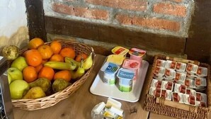 A wooden table with a basket of fruits, a glass of juice, and packaged snacks against a brick wall. - POSADA LA VICTORIA (Miengo)