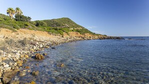 Un paysage côtier caractérisé par des eaux claires et peu profondes, un rivage rocheux et une colline couverte de végétation, sur laquelle se trouve un bâtiment.