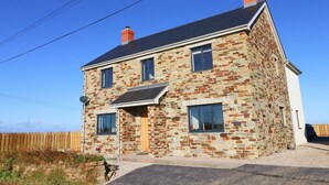 Une maison en pierre à deux étages, avec un toit à pignon, un porche à l’avant et une clôture en bois en arrière-plan.