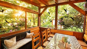 Interior de madera con un banco, una mesa y sillas, ventanas amplias que dan a árboles y flores.
