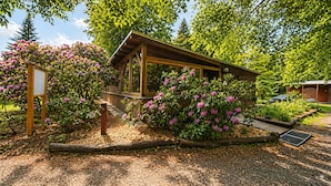 Una cabaña de madera con un porche cubierto, rodeada de arbustos y árboles en flor.