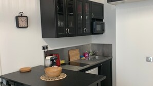 Eine kompakte Küche mit schwarzen Schränken, einer Mikrowelle und einer Kaffeemaschine. Ein Esstisch mit vier Stühlen und einer gewebten Tischmatte ist ebenfalls vorhanden.