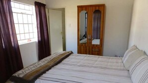 A bedroom with a bed, a wooden wardrobe, a mirror, and a window with curtains. - African Violet (Mbabane)