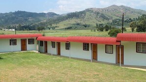 A row of simple, single-story buildings with red roofs, set against a backdrop of green hills and a clear sky. - African Violet (Mbabane)