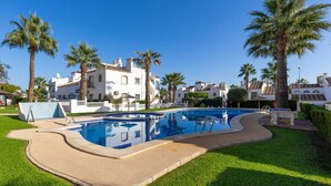 A swimming pool with a curved edge, surrounded by well-maintained grass and palm trees, with buildings in the background. - Infinity Haven by Fidalsa (Villamartín)