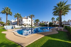 A swimming pool with a curved edge, surrounded by well-maintained grass and palm trees, with buildings in the background. - Infinity Haven by Fidalsa (Villamartín)