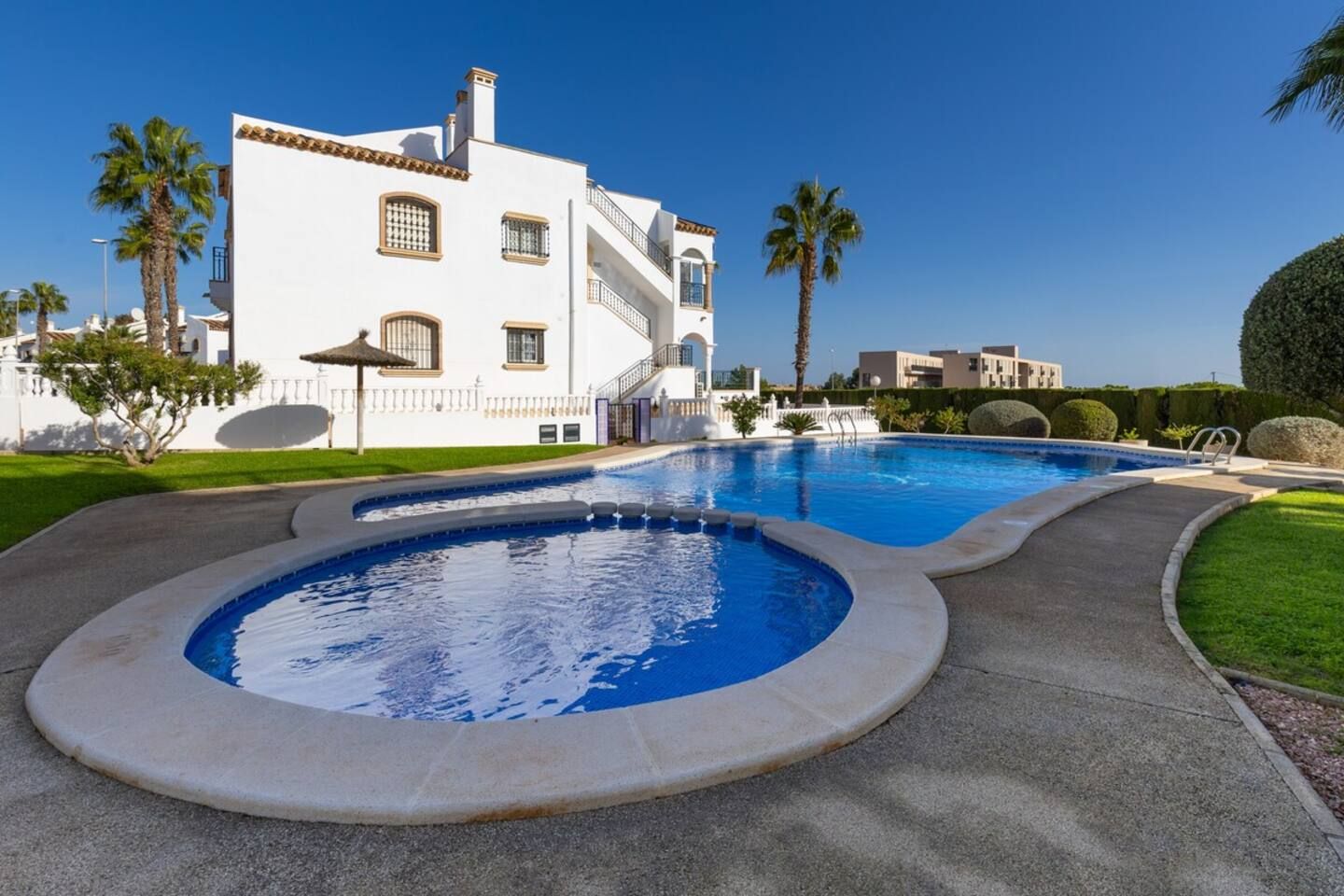 Une piscine au design incurvé, un bâtiment blanc doté de nombreuses fenêtres et de balcons, et une pelouse bien entretenue agrémentée de palmiers.
