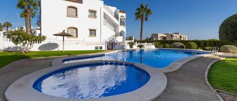 A swimming pool with a curved design, a white building with multiple windows and balconies, and a well-maintained lawn with palm trees.
