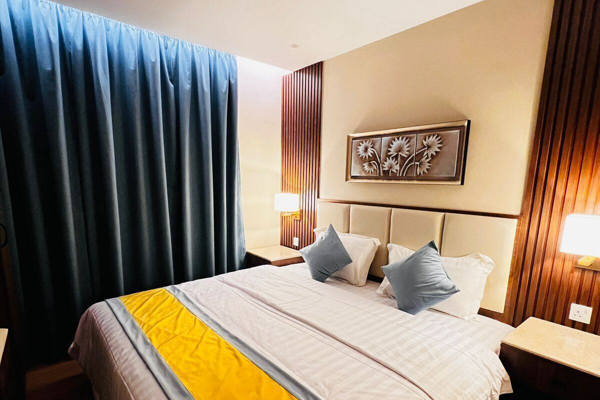 Una habitación de hotel con una cama grande, un cabecero, una obra de arte colgada en la pared, una mesita de noche con una lámpara y una ventana con cortinas.