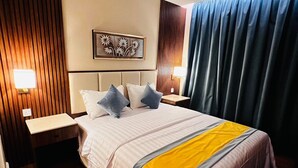 A hotel room with a large bed, wooden panel walls, a ceiling light, a wall-mounted artwork, and a window with curtains. - Pestana Apartment 4 (Riyadh)