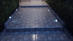 A paved pathway leading to a white door, flanked by hedges and a chair. - Elite Apartment Larisa (Larissa)