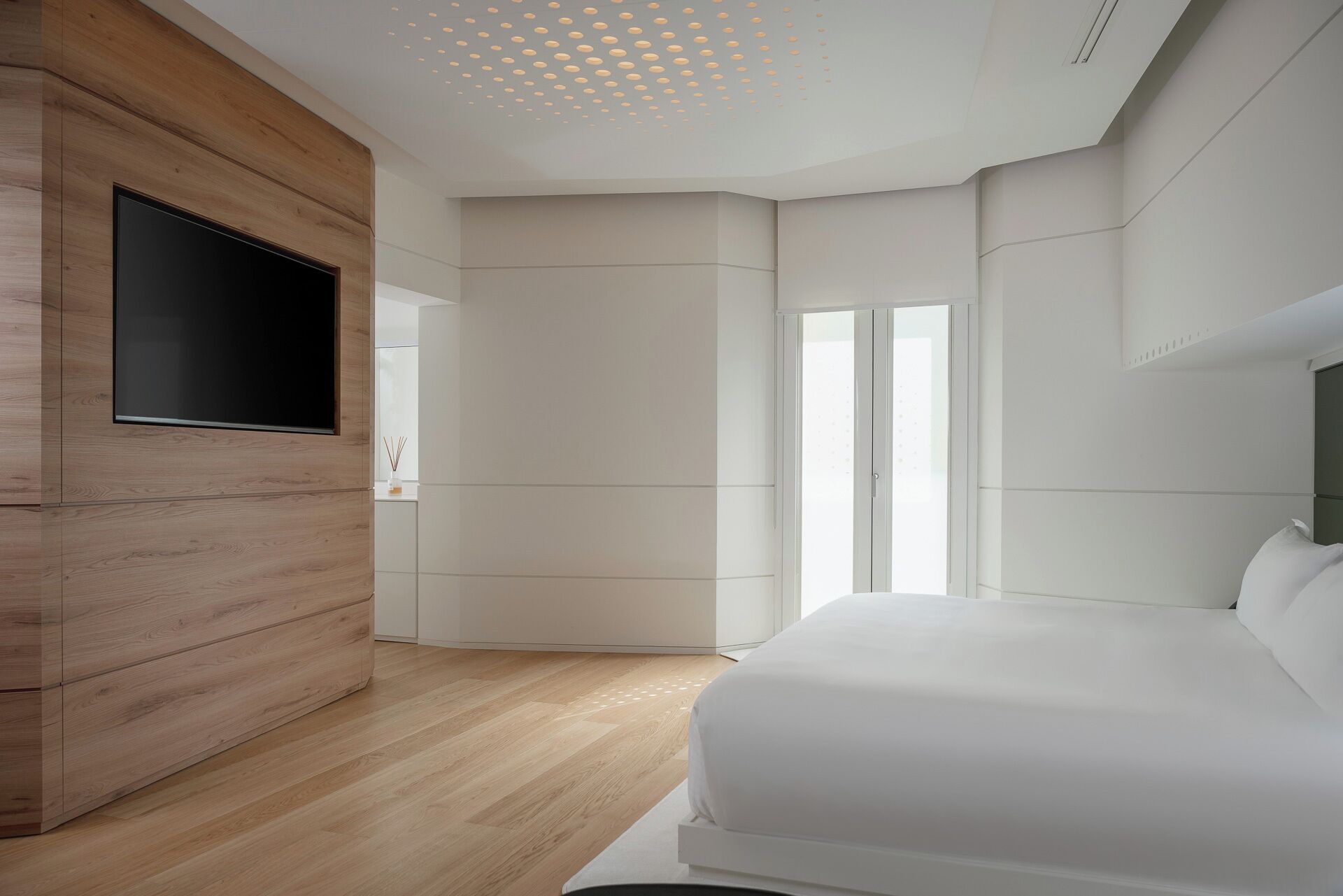 A modern bedroom with a wooden wardrobe, a flat-screen TV, a large bed, and a sliding door leading to a balcony.