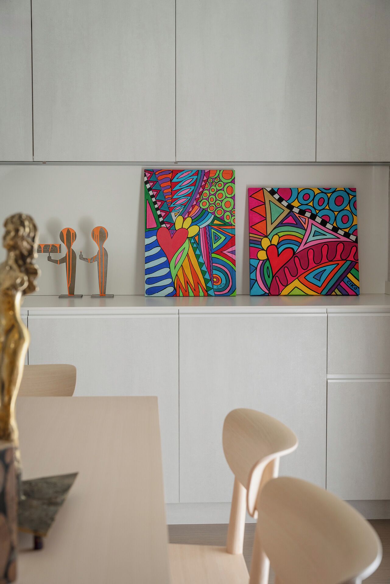 A modern kitchen with white cabinets, two colorful abstract paintings, and a dining area with a wooden table and chairs.
