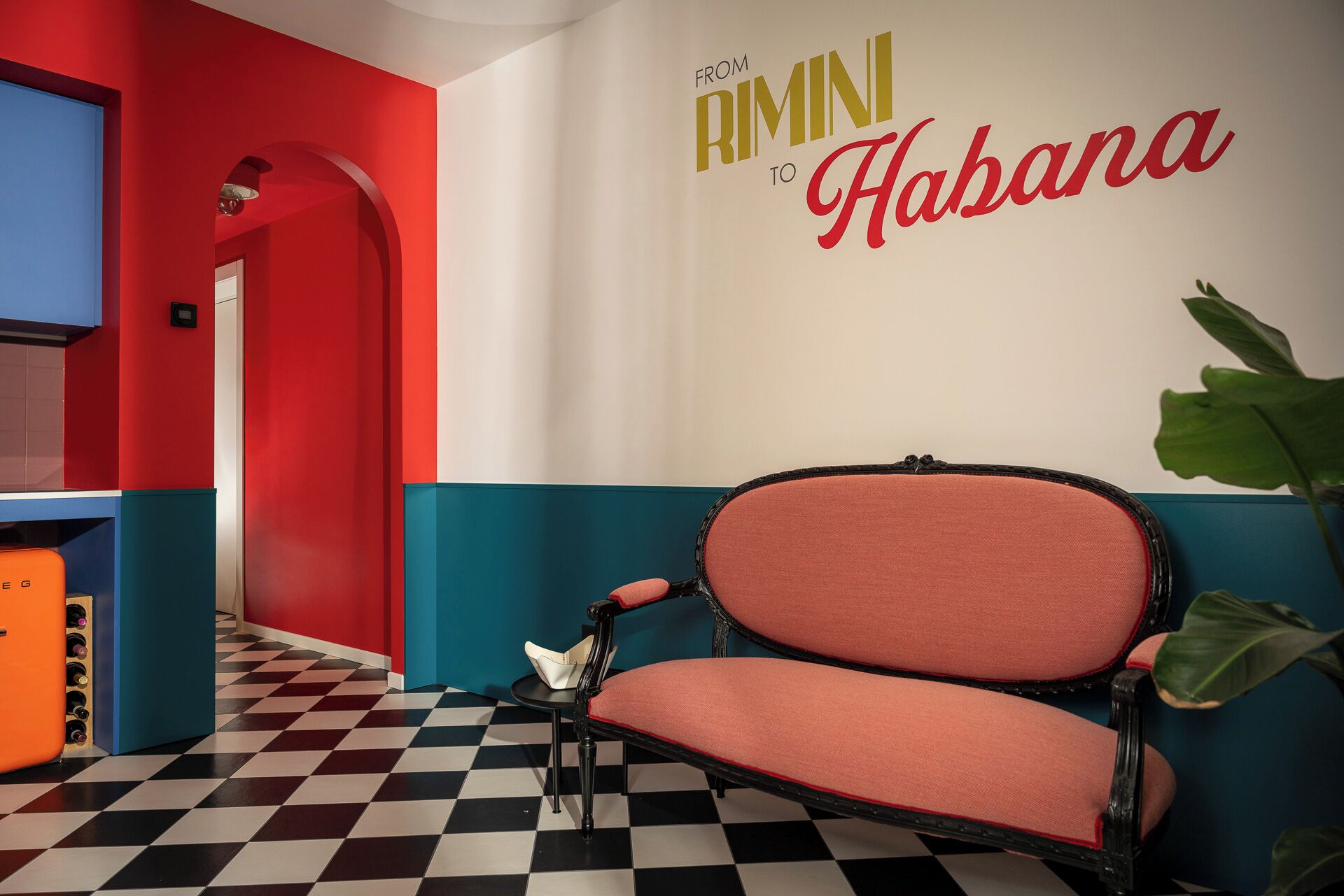 Et skakmønstret gulv, en væg i røde og turkise farver, en vintage stol og et skilt på væggen med teksten 'FROM RIMINI TO HABANA'.