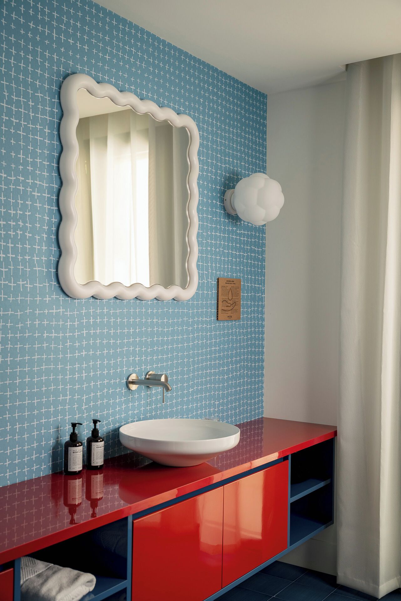 A bathroom with a blue patterned wall, a white sink, red cabinets, and a white shower curtain.