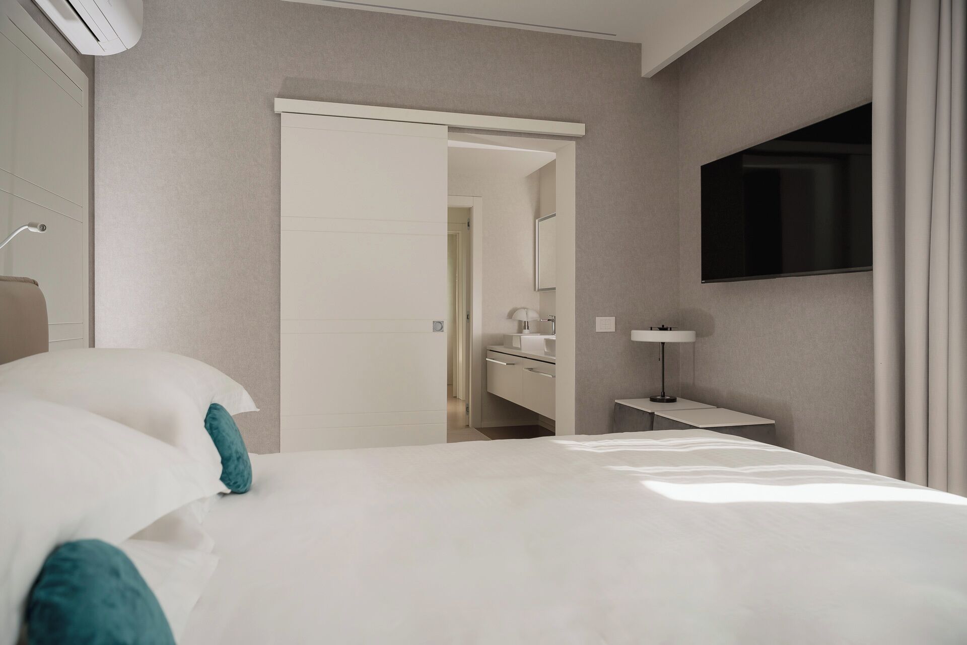 A modern hotel room with a large bed, a bedside table with a lamp, a TV mounted on the wall, and a bathroom visible through an open door.