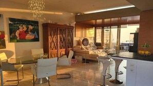 A modern living room with a glass dining table, white chairs, a sofa, and a large painting on the wall. - Exclusive Duplex Penthouse with Jacuzzi and Panoramic Views in Valletta  (Sliema)