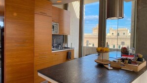 A modern kitchen with wooden cabinets, a built-in oven, and a large window offering a city view. - Exclusive Duplex Penthouse with Jacuzzi and Panoramic Views in Valletta  (Sliema)