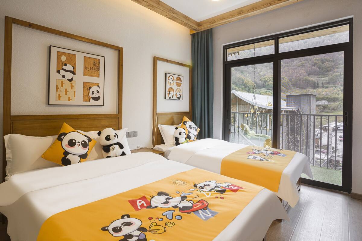 Habitación de hotel con dos camas, ropa de cama con temática de panda, cuadros en las paredes y un balcón con vistas al exterior.