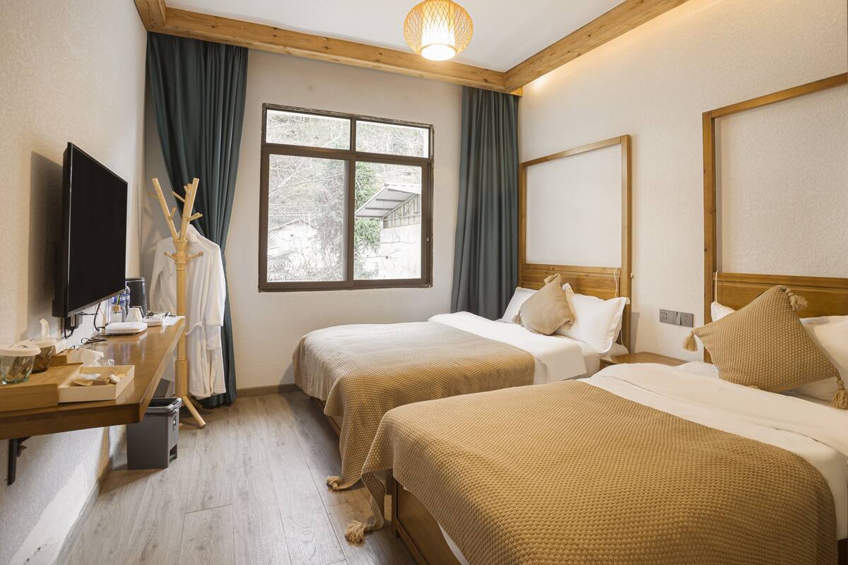Habitación de hotel con dos camas, televisión, escritorio con lámpara y ventana con cortinas.