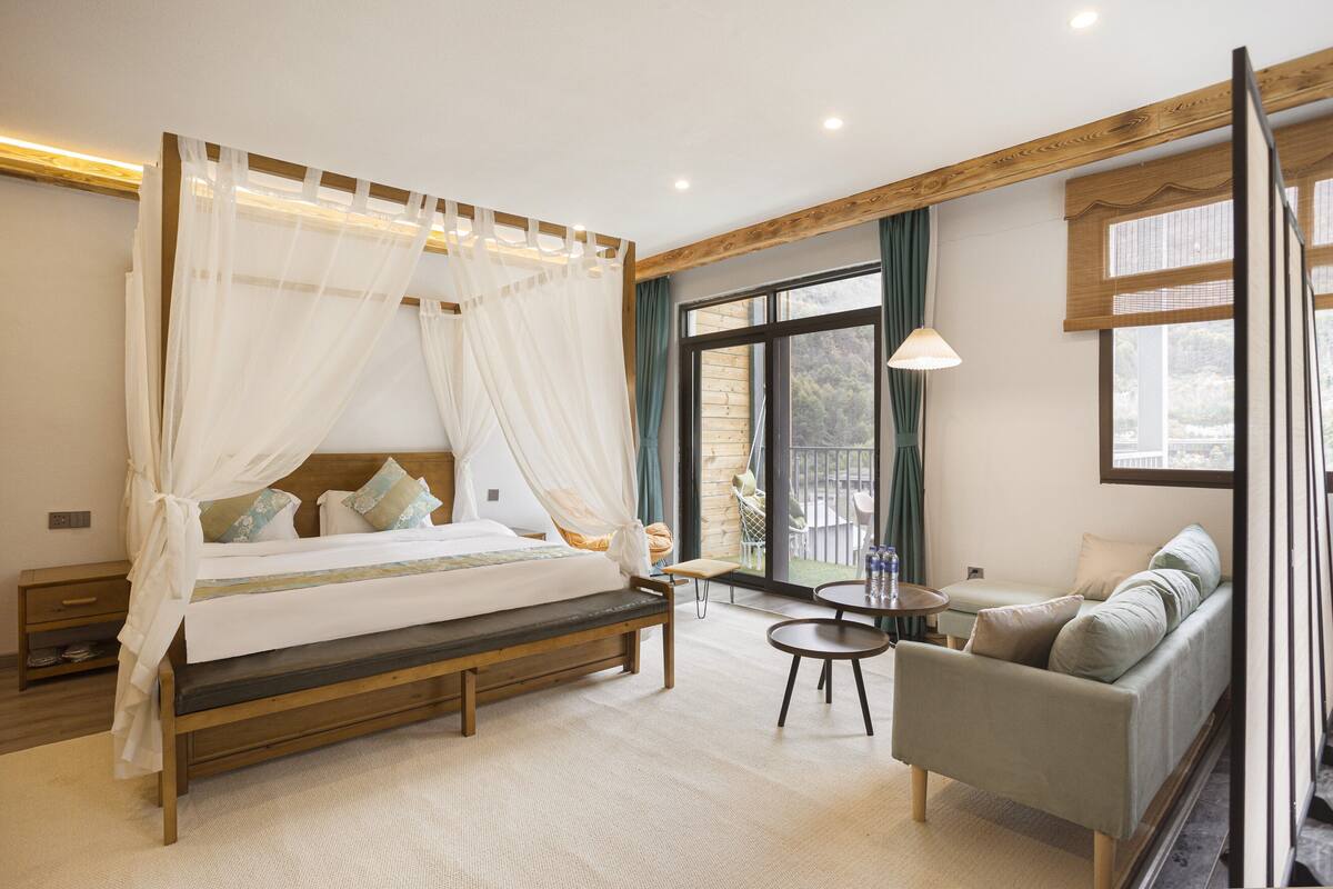 Un dormitorio con una cama con dosel, un sofá, una mesita redonda y vistas al exterior a través de un ventanal corredero.
