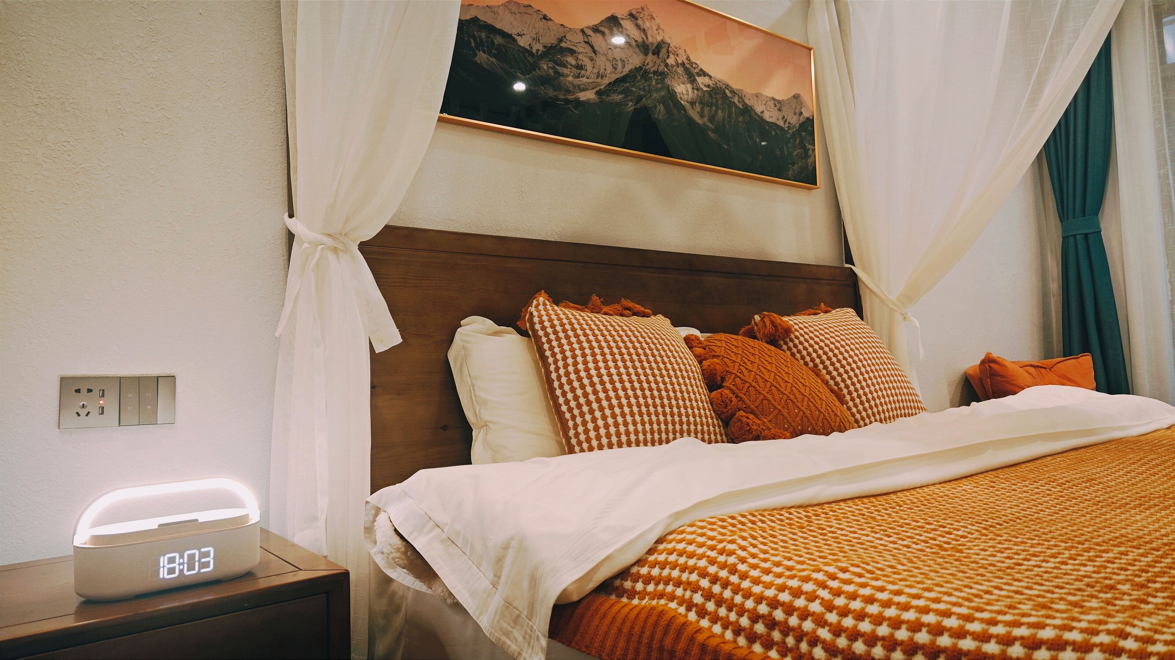 Una cama bien hecha con almohadas estampadas, una mesita de noche con un reloj digital y un cuadro de un paisaje montañoso sobre la cama.