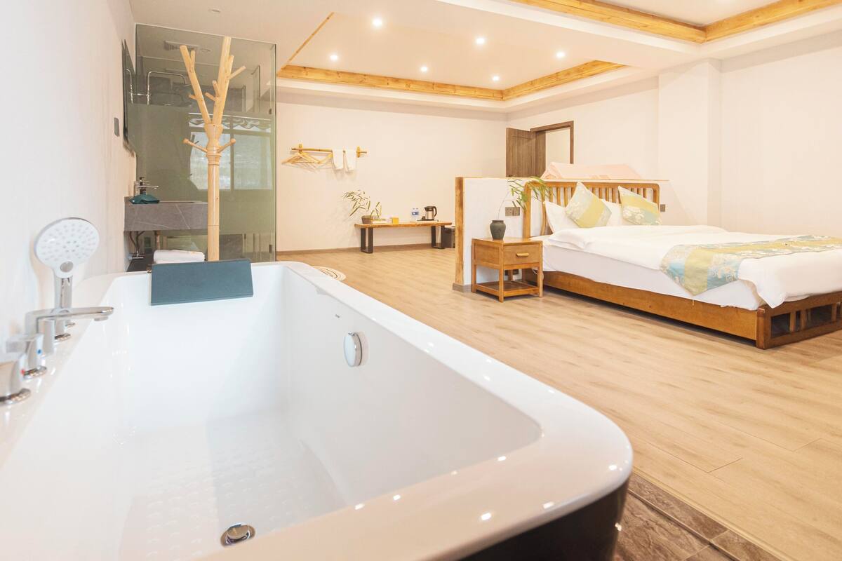 Un dormitorio moderno con una bañera grande, piso de madera, una cama con cabecera de madera y una mesita de noche con lámpara.