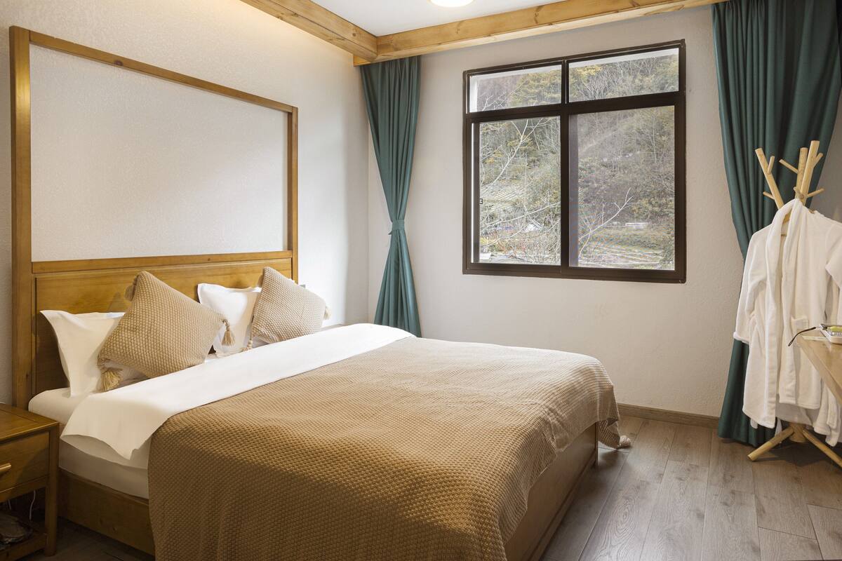 Una cama bien hecha con cabecera de madera, una ventana con cortinas y una mesita de noche de madera con lámpara.