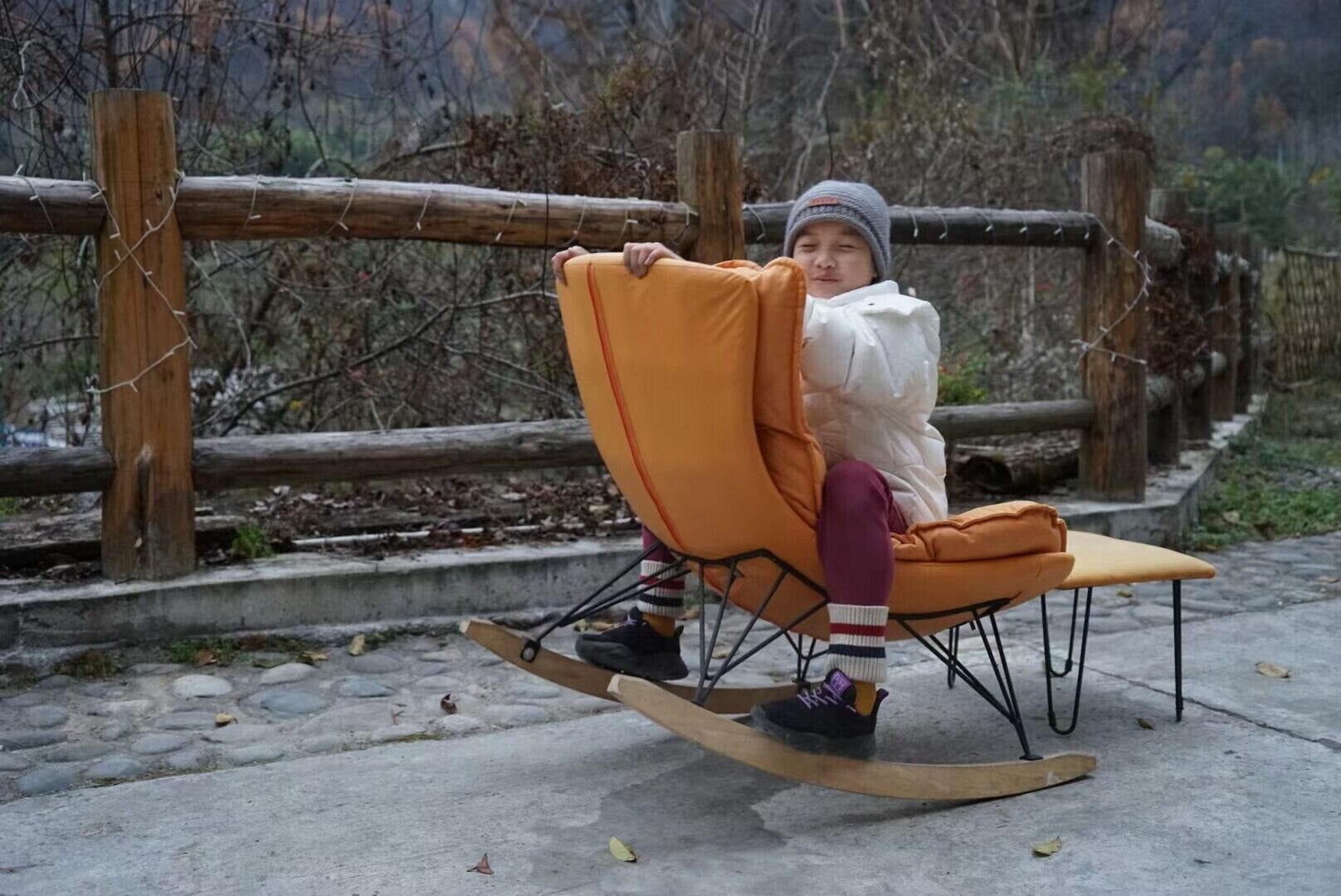 A person sitting on a rocking chair with a wooden base and orange cushions, outdoors with a wooden fence and trees in the background.