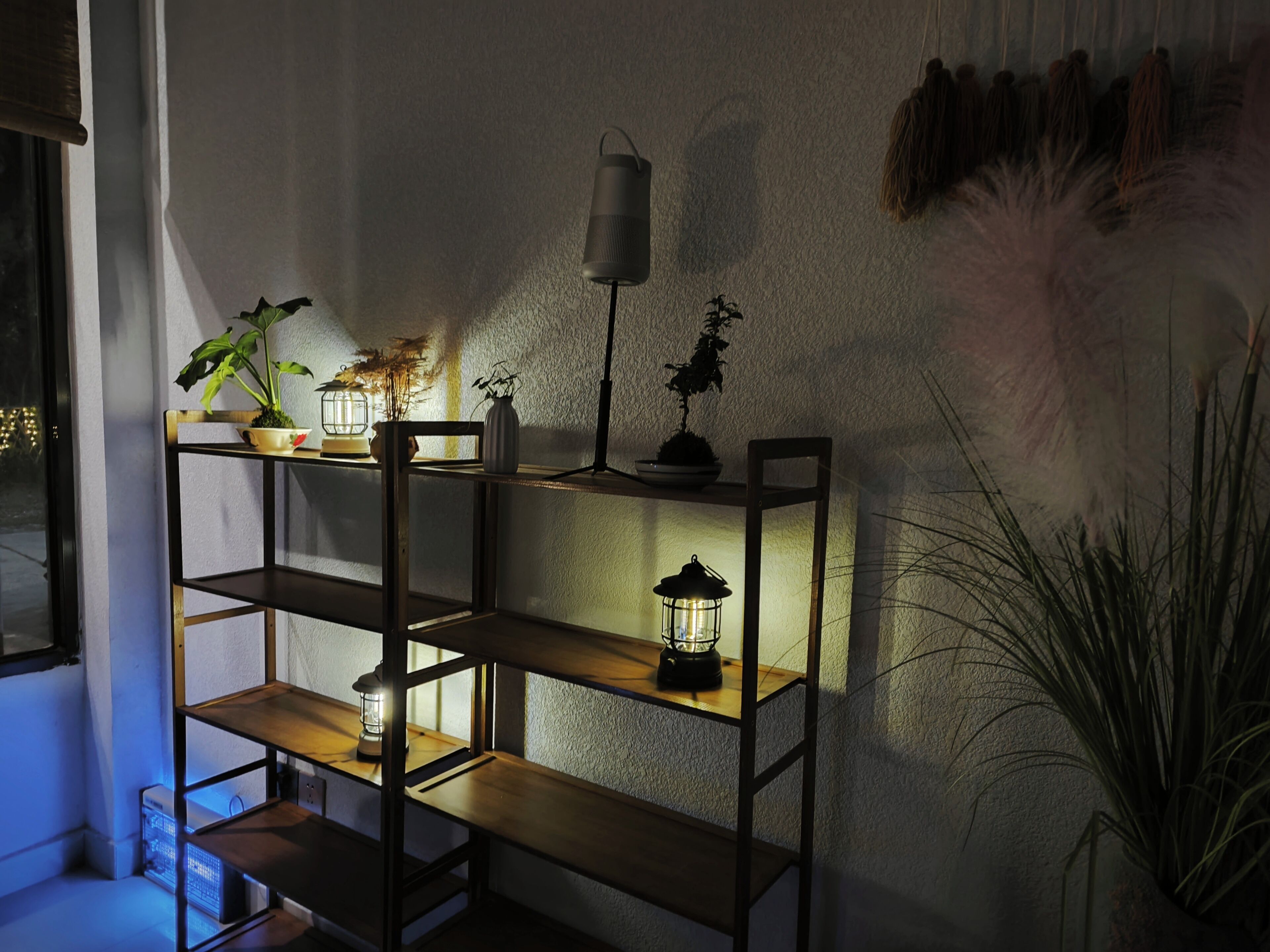 A wooden shelf unit with multiple shelves, each holding a lit lantern. There are potted plants on the shelves, and a wall-mounted lantern above.