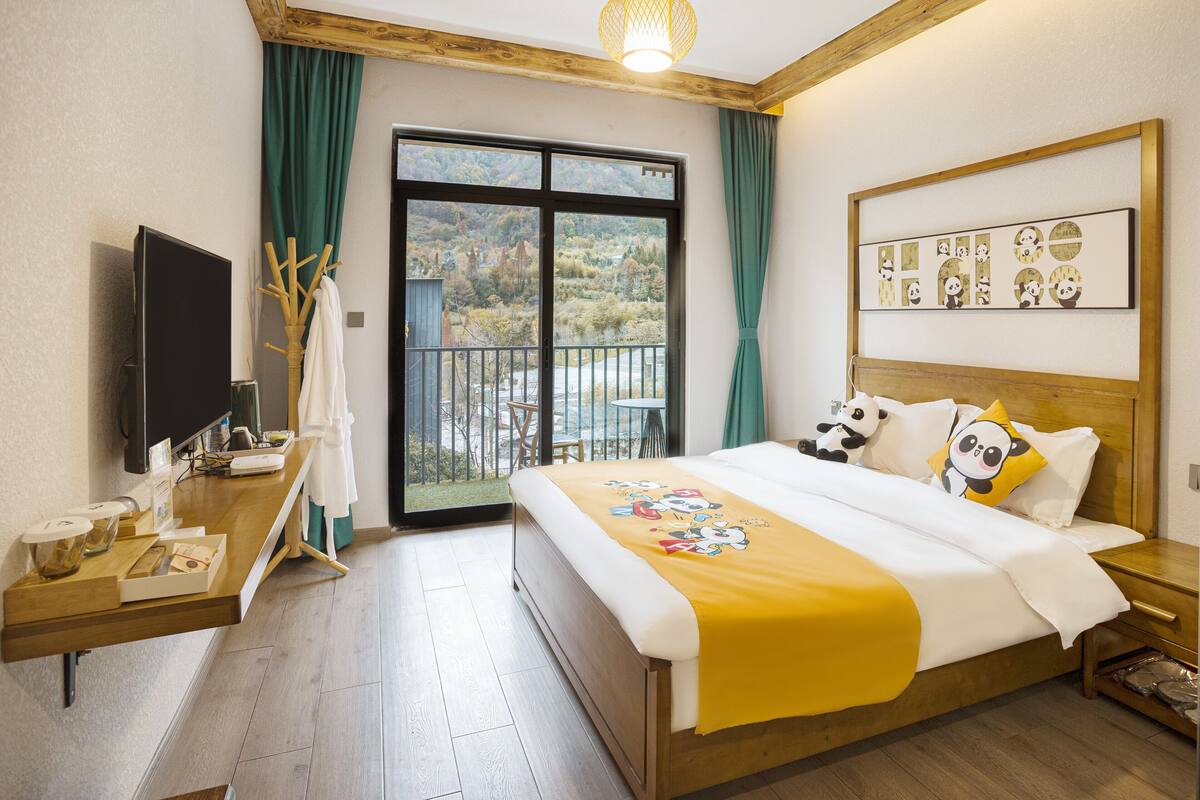 Habitación de hotel con cama, televisor, escritorio de madera, balcón con vistas a las montañas y ventana con cortinas.