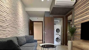 A modern living room with a grey sofa, a round black coffee table, a hanging light fixture, a washing machine, and a TV mounted on a wooden wall. - Saigon Royal View Residence & River (Ho Chi Minh City)