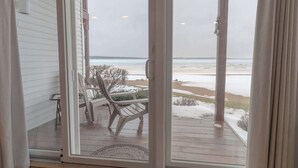 On aperçoit, à travers une porte-fenêtre coulissante, une terrasse en bois avec une chaise et une table, d’où l’on a une vue sur une plage enneigée.