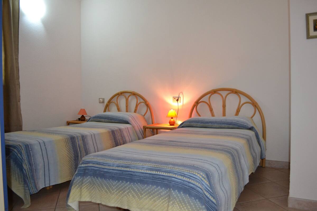 A room with two single beds, each with a headboard, a bedside table with a lamp, and a wall-mounted light fixture.