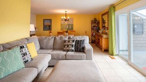 A living room with a grey sofa, patterned cushions, a chandelier, and a dining area with a table and chairs.
